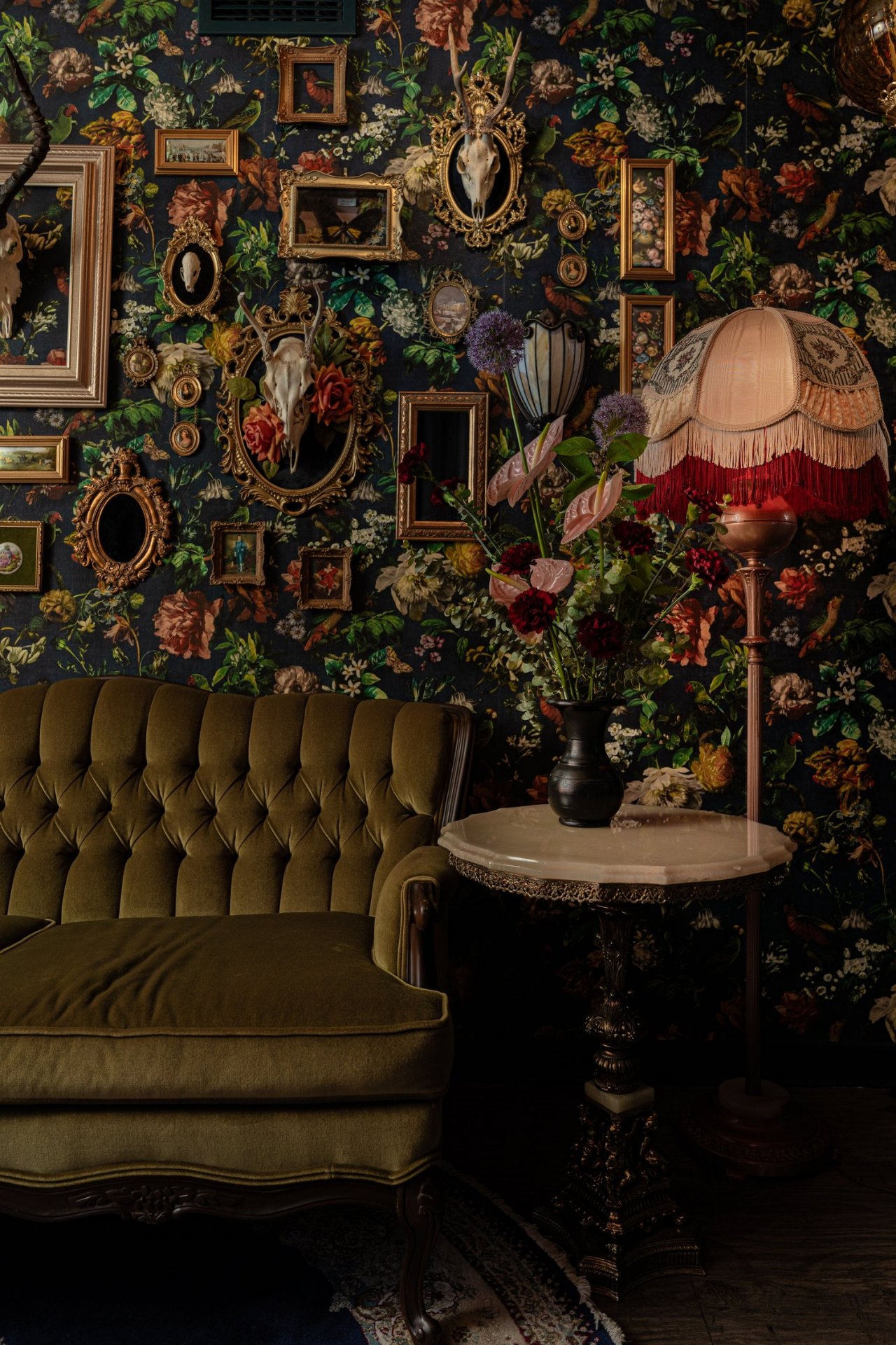 Glitterbox Studio interior — olive velvet sofa, ornate side table, and floral gallery wall.