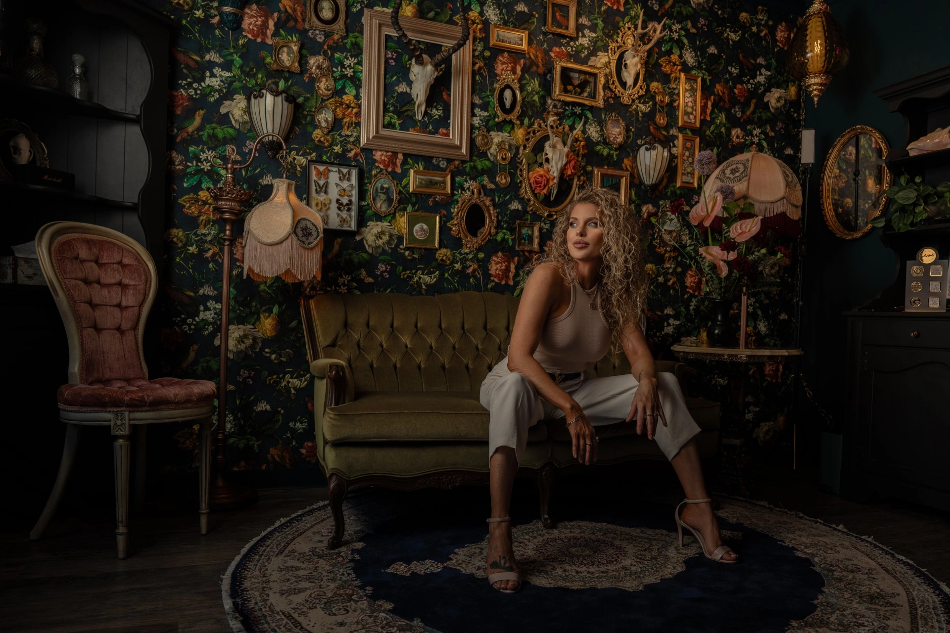 Rhiana Lee seated in Glitterbox Studio — vintage olive sofa against a bold floral gallery wall.